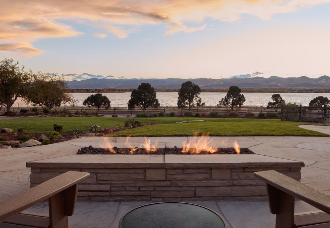 Scenic outdoor fire pit with lake and mountain view at sunset in a landscaped garden.
