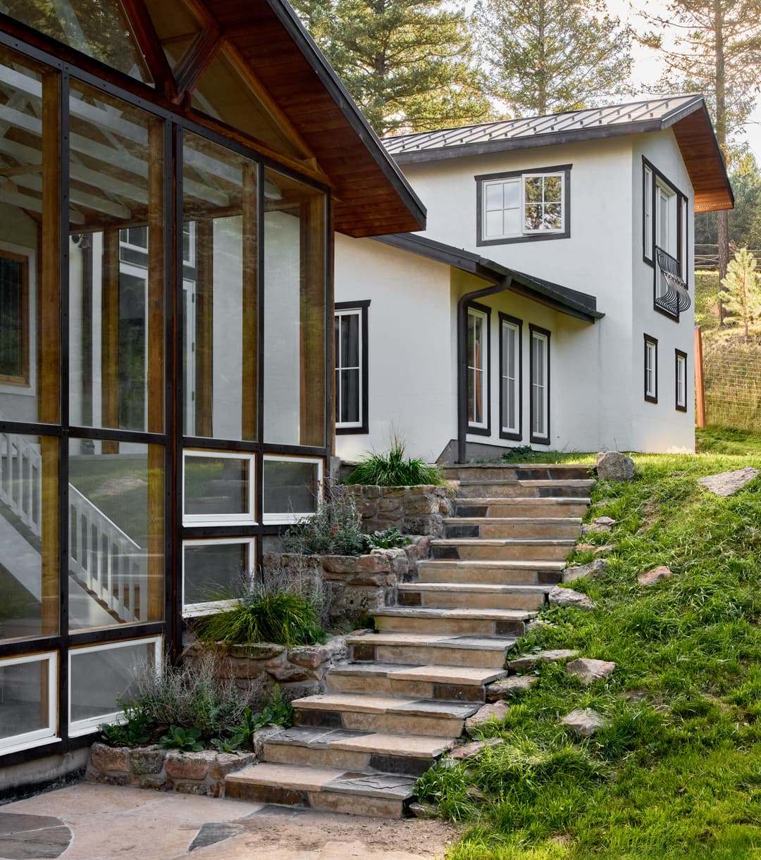 Modern house exterior with glass wall, stone steps, and lush greenery in a natural setting.