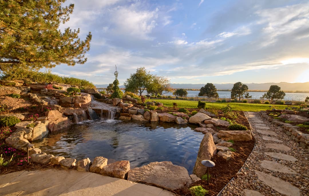 Scenic garden with a pond, stone pathway, and sunset view over a lake.