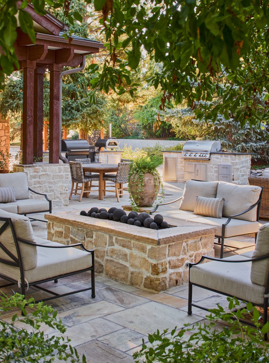 Patio with stone fire pit, outdoor seating, grill area, and lush greenery in a backyard setting.