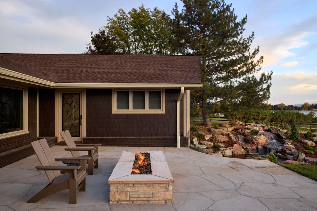 Modern outdoor patio with fire pit, seating, and lush landscaping by a serene lake.