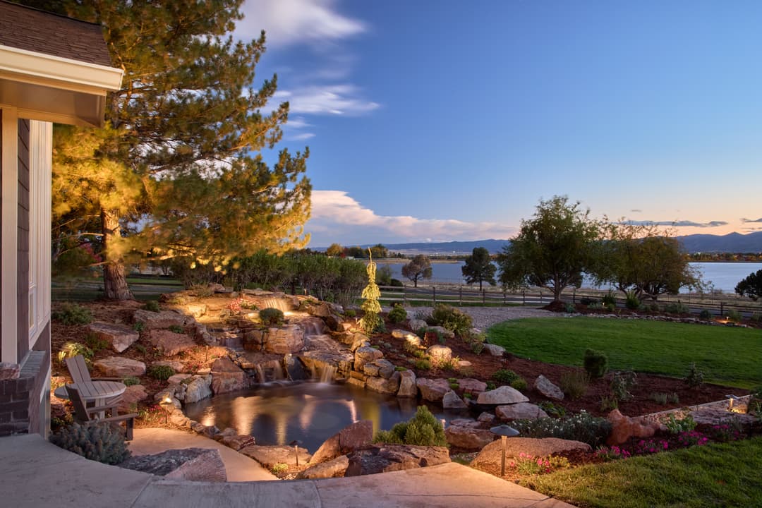 Serene backyard landscape with a pond, surrounded by greenery and lit by warm evening lights.