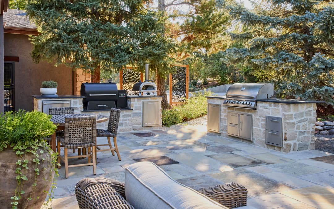 Outdoor kitchen with grills, stone patio, dining area, and lush greenery in a serene garden setting.
