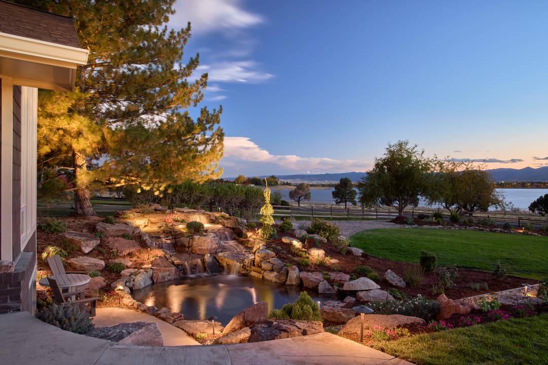 Scenic backyard with illuminated pond, rocks, and lush landscaping at sunset.