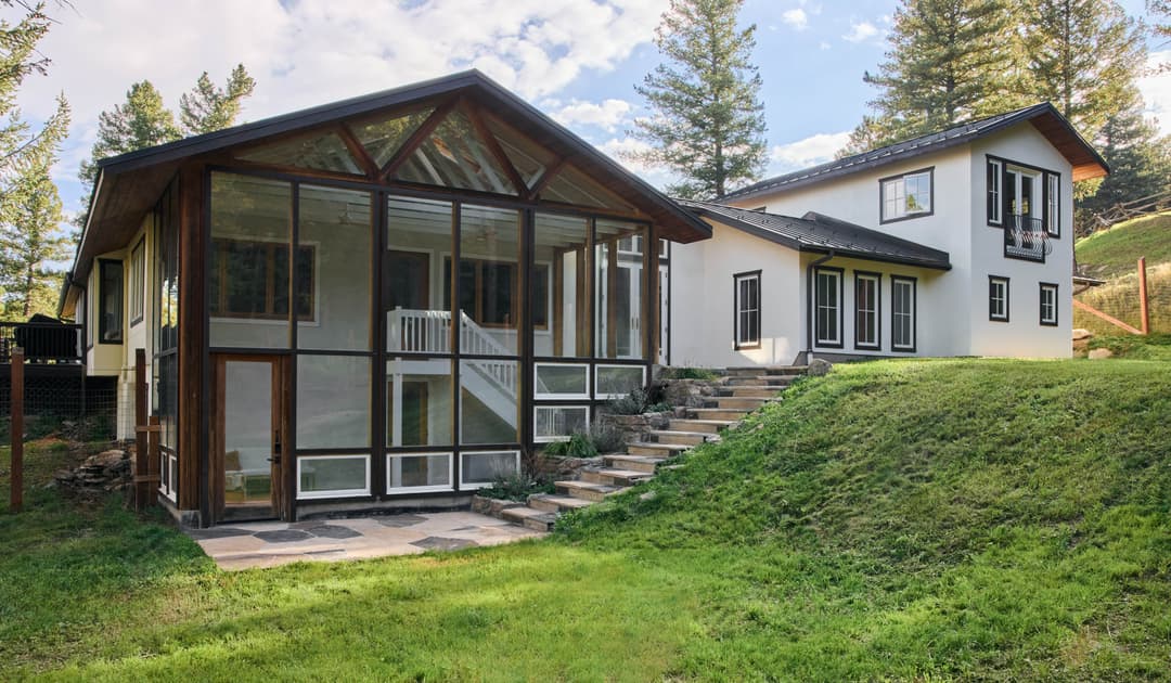Modern home with glass facade and landscaped yard, surrounded by tall trees and blue skies.