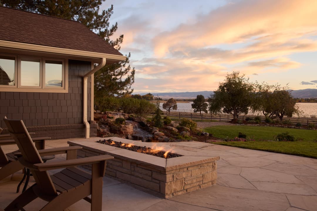 Cozy outdoor patio with stone fire pit overlooking a serene lake at sunset.