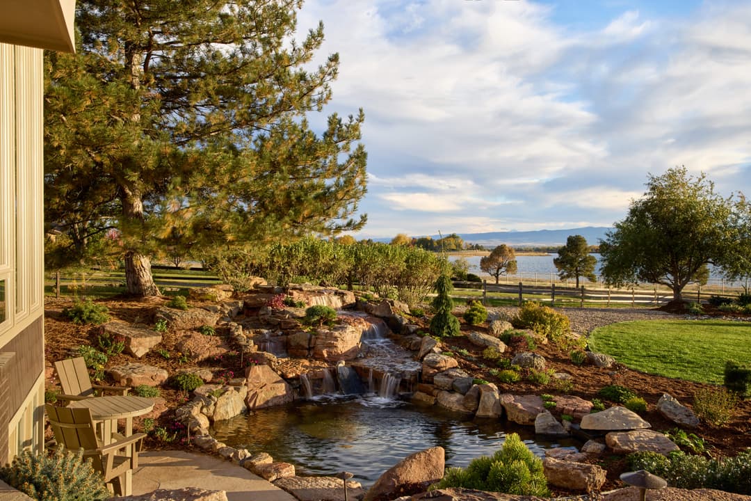 Lush garden with a pond and waterfall by a lake, surrounded by trees and scenic views.