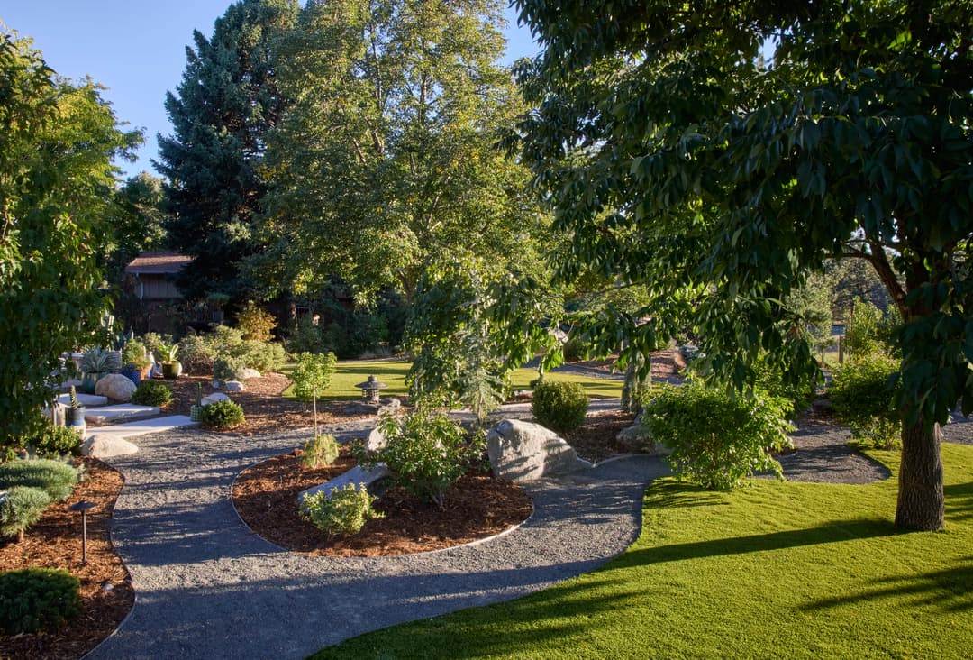 Lush garden with winding paths, trees, and decorative rocks under a clear blue sky.