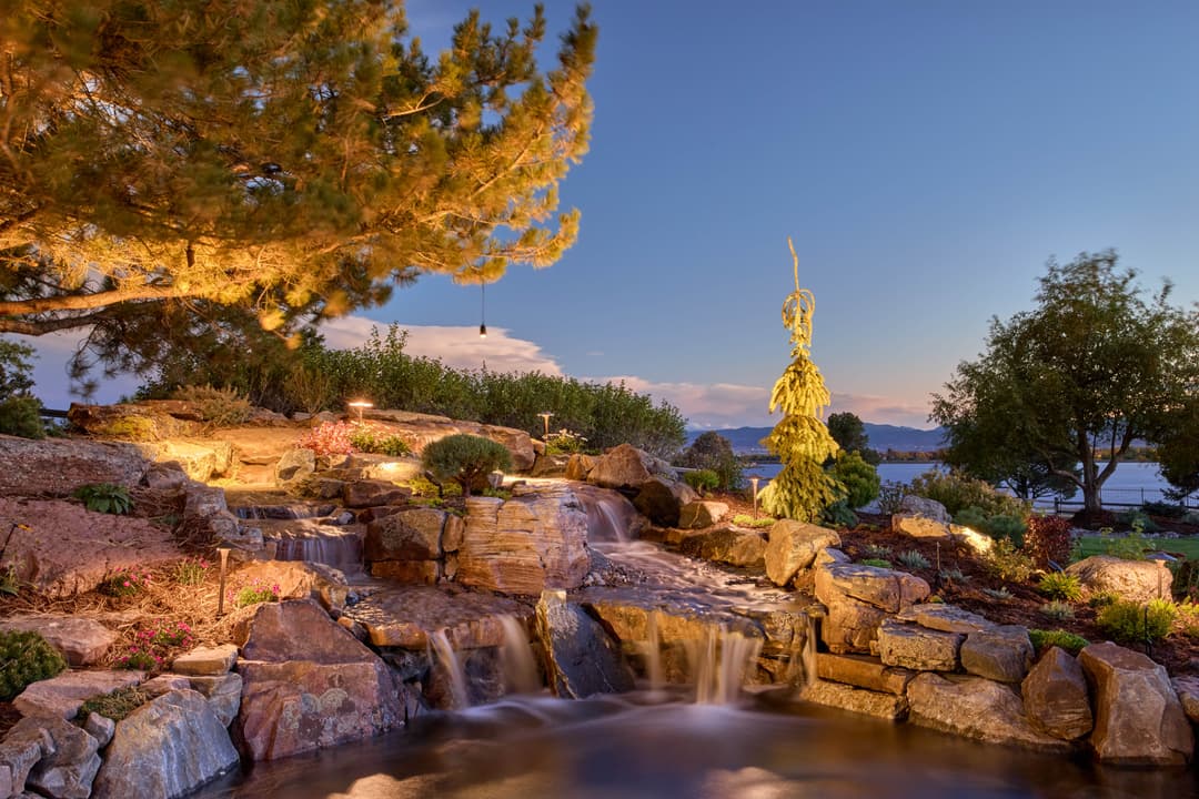 Serene evening garden with a flowing waterfall, rocky landscape, and soft lighting by the lake.
