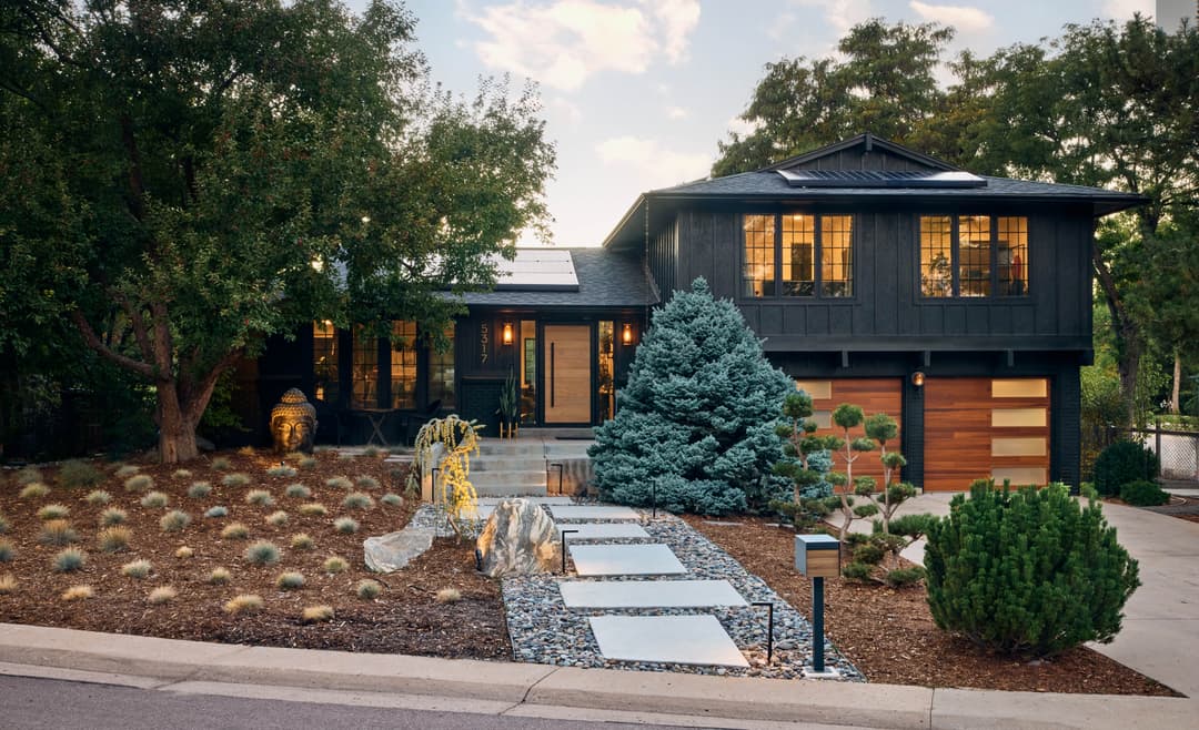 Modern black house with landscaped garden, stone pathway, and solar panels in a serene setting.