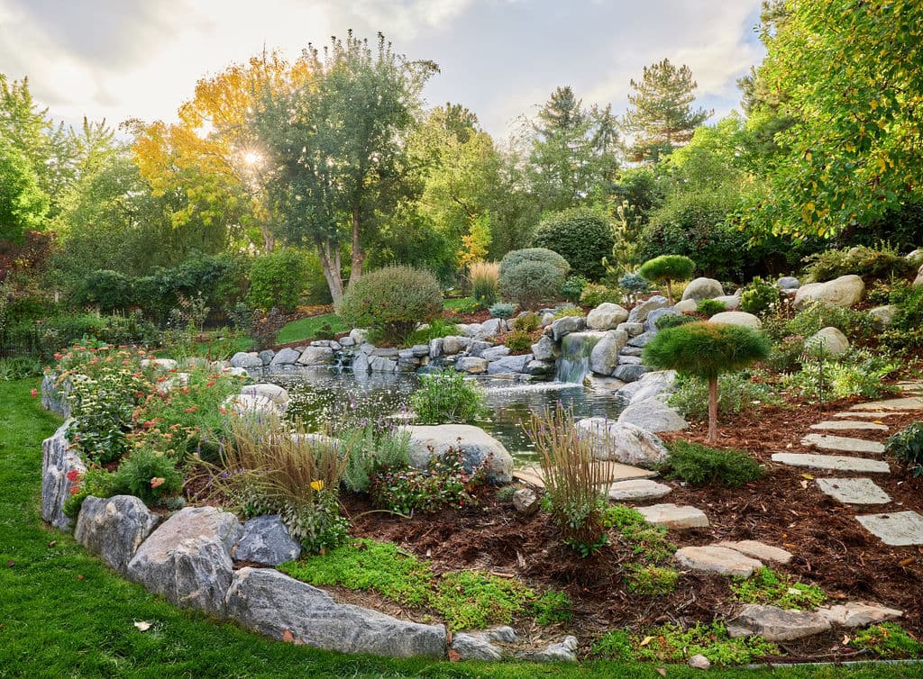 Lush garden landscape with a pond, waterfall, stepping stones, and vibrant flowers at sunset.