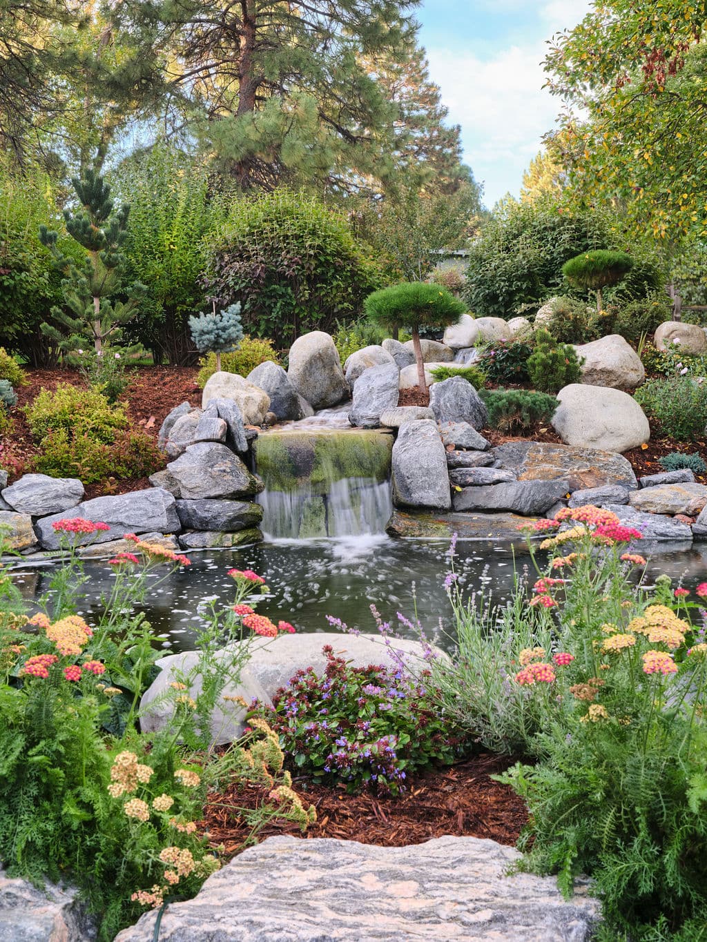 Serene garden pond with a waterfall surrounded by vibrant flowers and lush greenery.