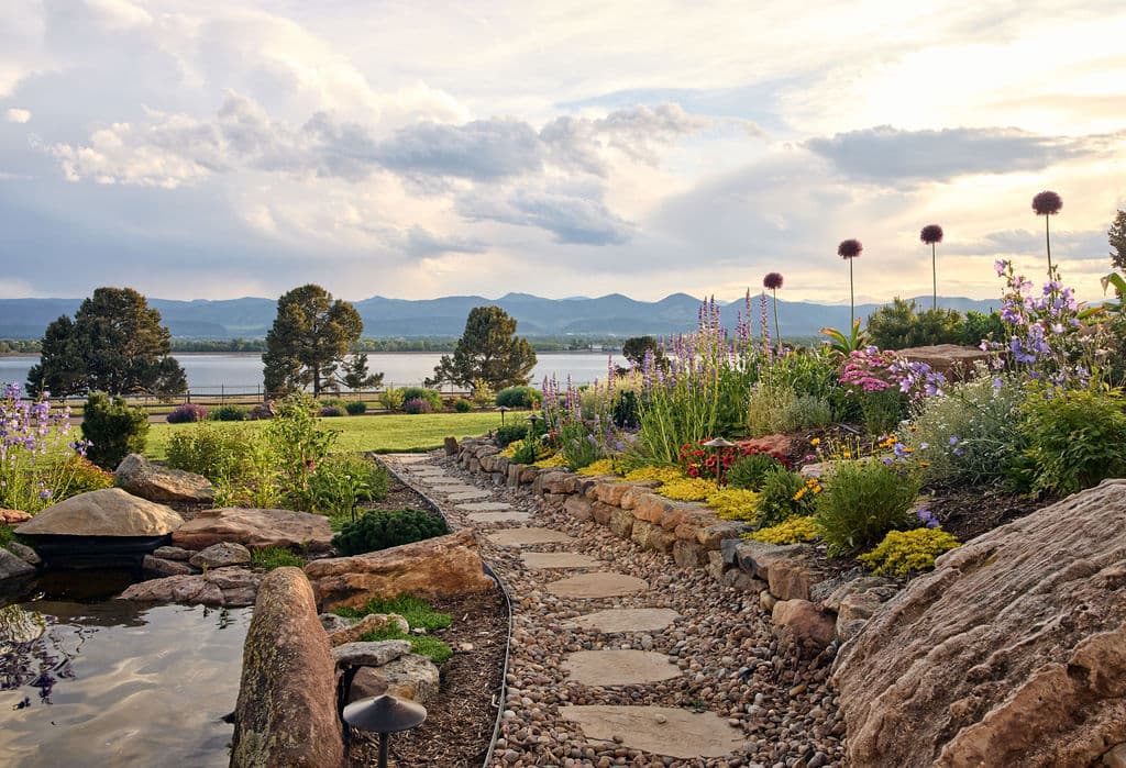 Colorful garden path leading to a lake with mountains in the background at sunset.