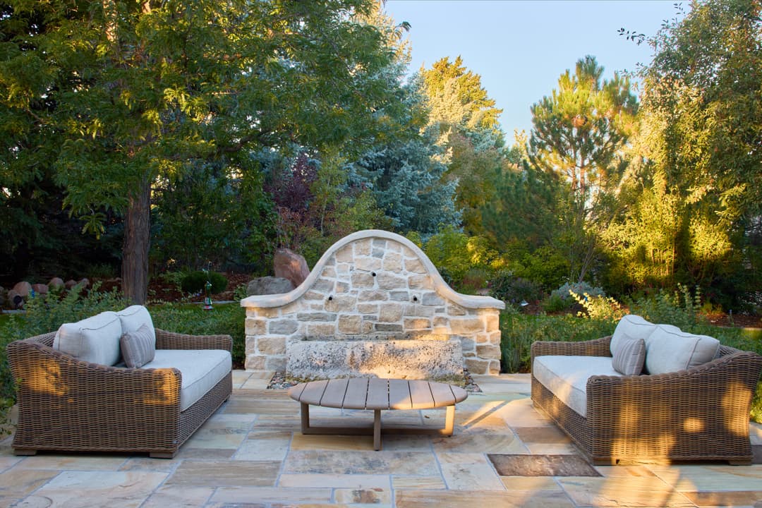 Cozy outdoor seating area with stone wall and lush greenery in sunlight. Perfect for relaxation.