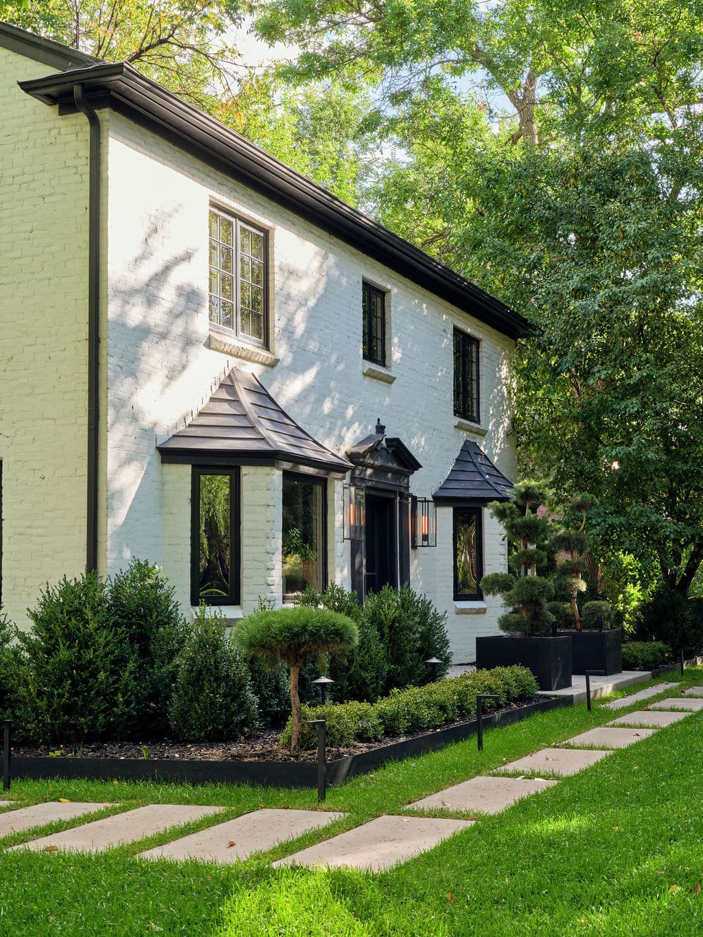 Stylish white brick house with black accents, surrounded by lush greenery and a path.