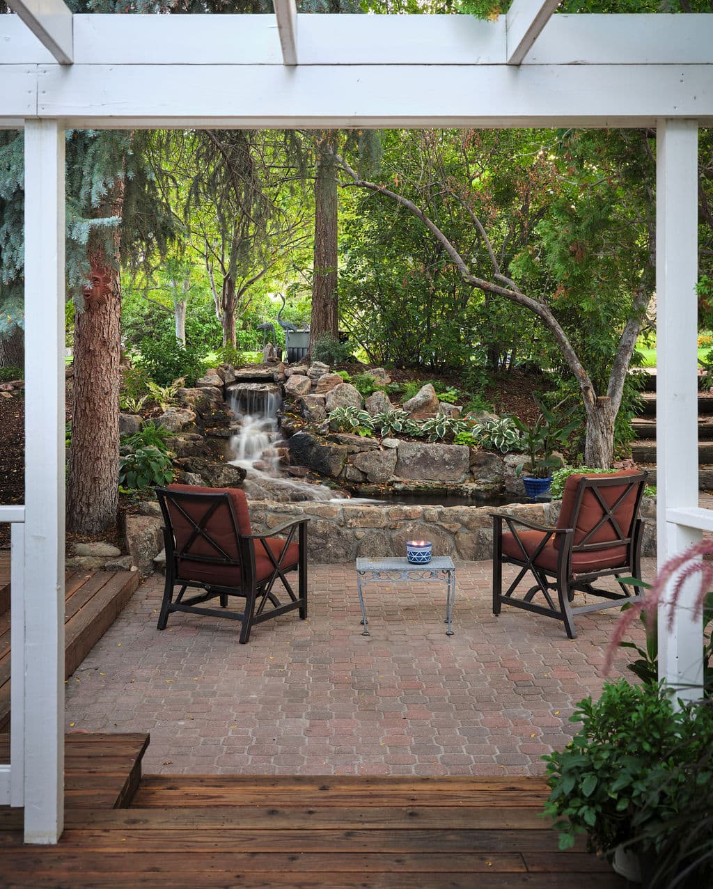 Serene backyard patio with chairs, stone waterfall, and lush greenery in a tranquil setting.