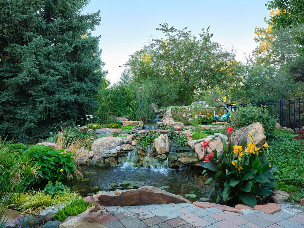 Lush garden with a serene pond, waterfall, and vibrant flowers surrounded by trees.