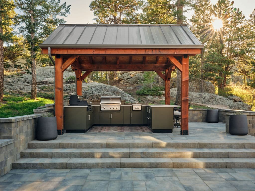 Outdoor kitchen under wooden gazebo, featuring a grill and stone patio in a scenic natural setting.