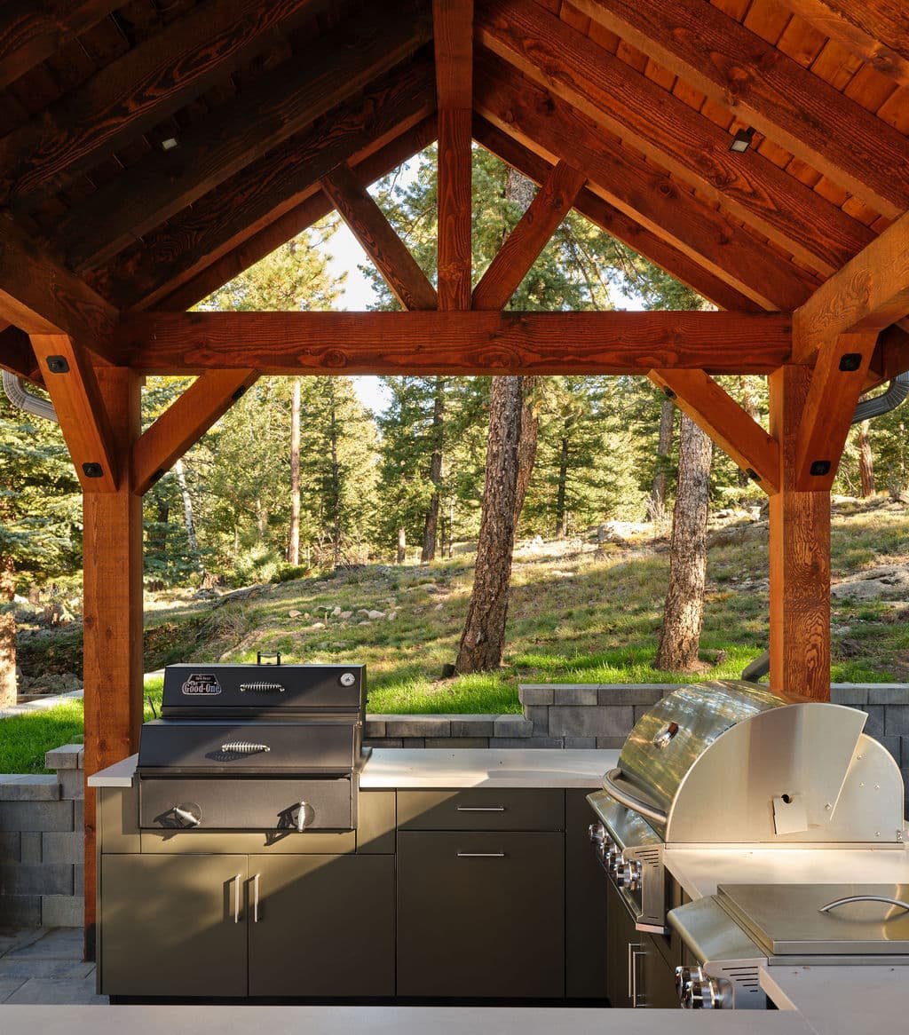 Outdoor kitchen under a rustic wooden pavilion with grill and countertop in a forest setting.
