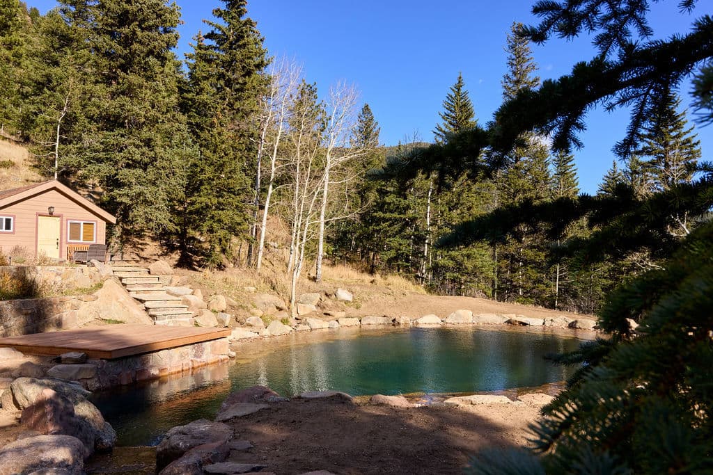 Scenic natural hot springs with a wooden deck, surrounded by pine trees and mountains.