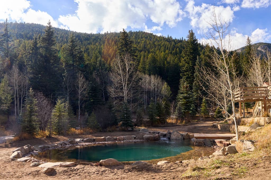 Natural hot spring surrounded by pine trees and mountains under a partly cloudy sky.