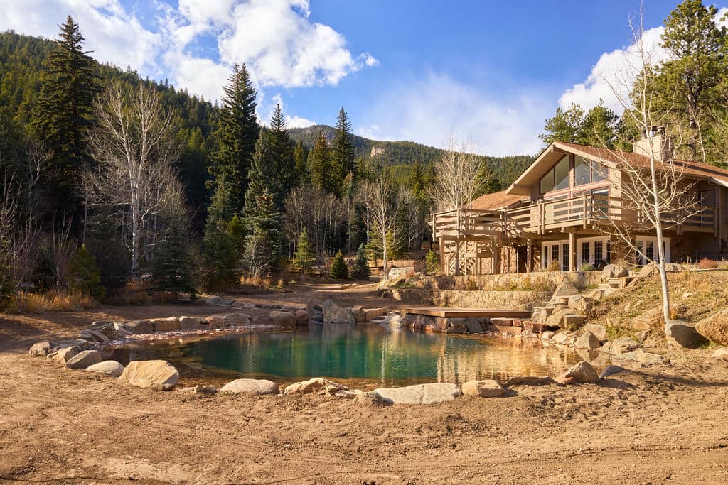 Scenic cabin near a tranquil pond surrounded by mountains and trees in bright sunlight.