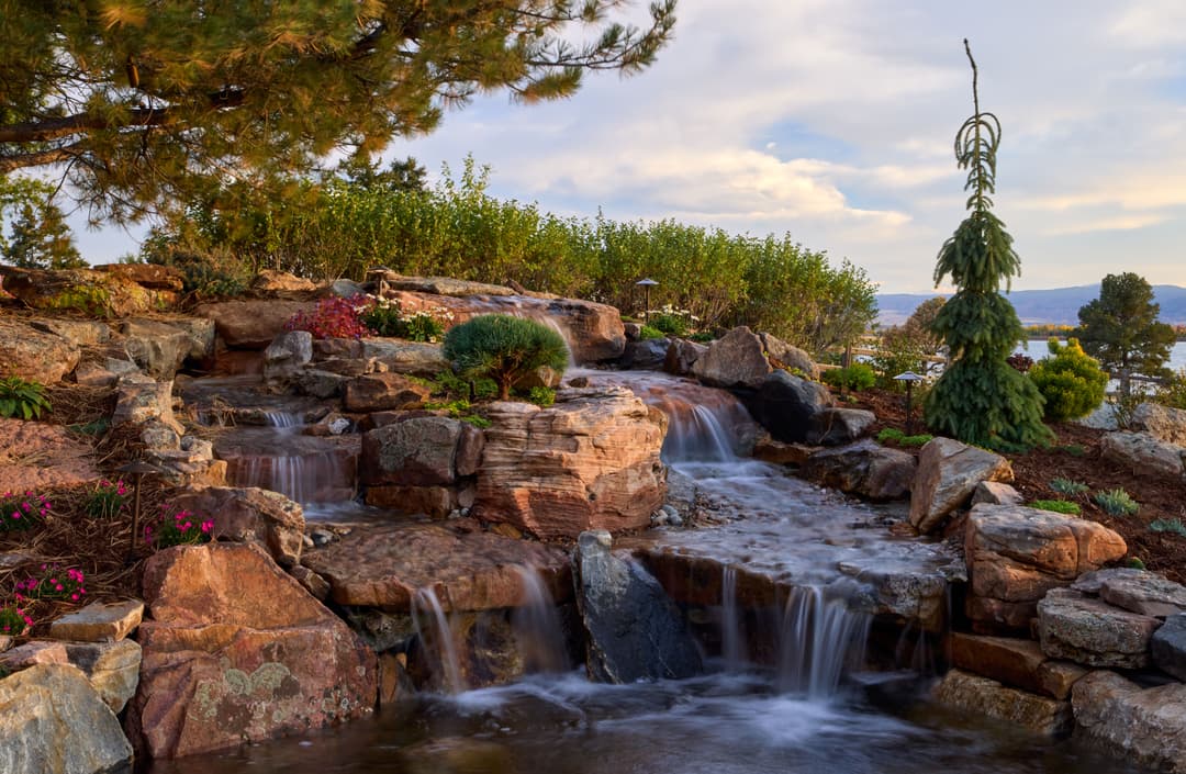 Lush garden waterfall with stones, greenery, and a scenic lake view in the background.