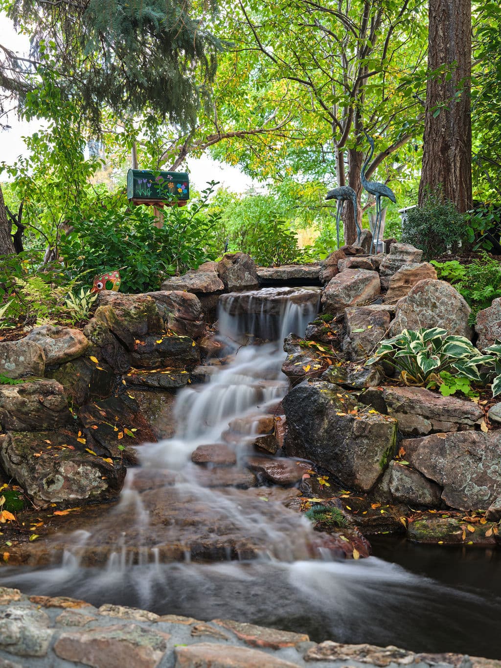 Tranquil garden waterfall cascading over rocks, surrounded by vibrant greenery and trees.