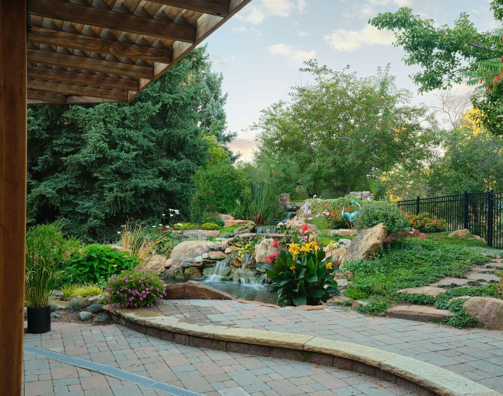 Lush garden with colorful flowers, cascading water feature, and stone pathway under a wooden pergola.