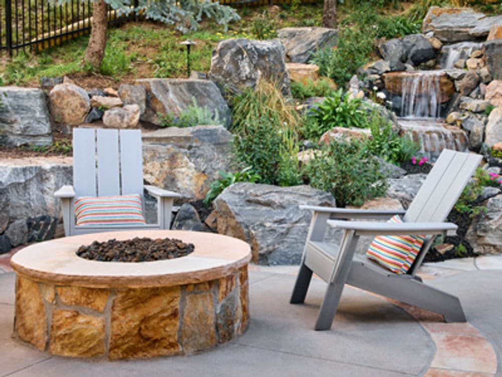 Cozy outdoor seating area with two gray chairs around a stone fire pit and lush greenery.