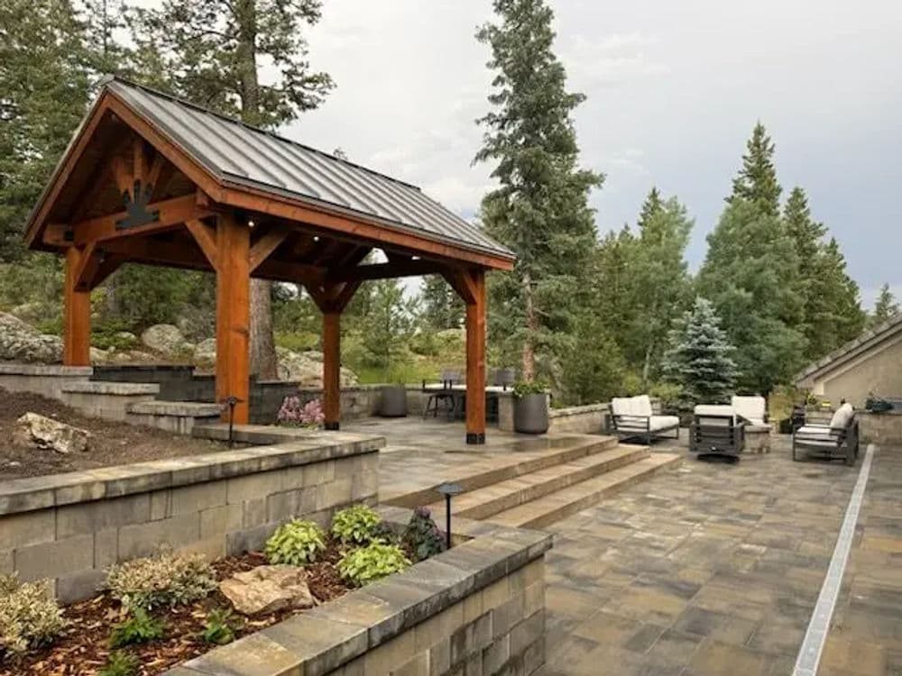 Wooden gazebo in landscaped garden with stone patio and seating area surrounded by trees.